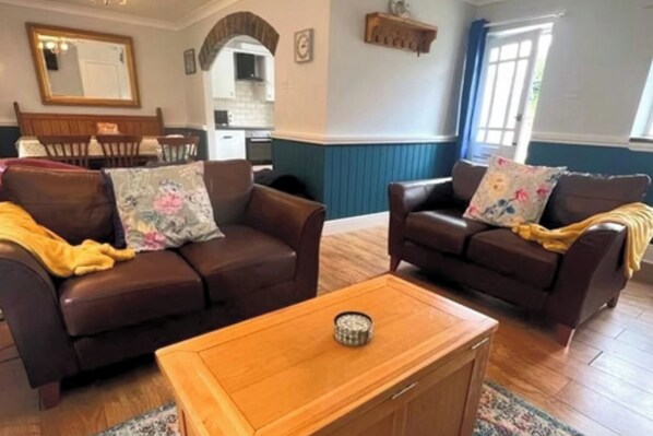 A living room with two brown leather sofas, a wooden coffee table, and a kitchen in the background. - 3 Bed Cottage Sleeps 6 - Dog Friendly - Patio (New Quay)