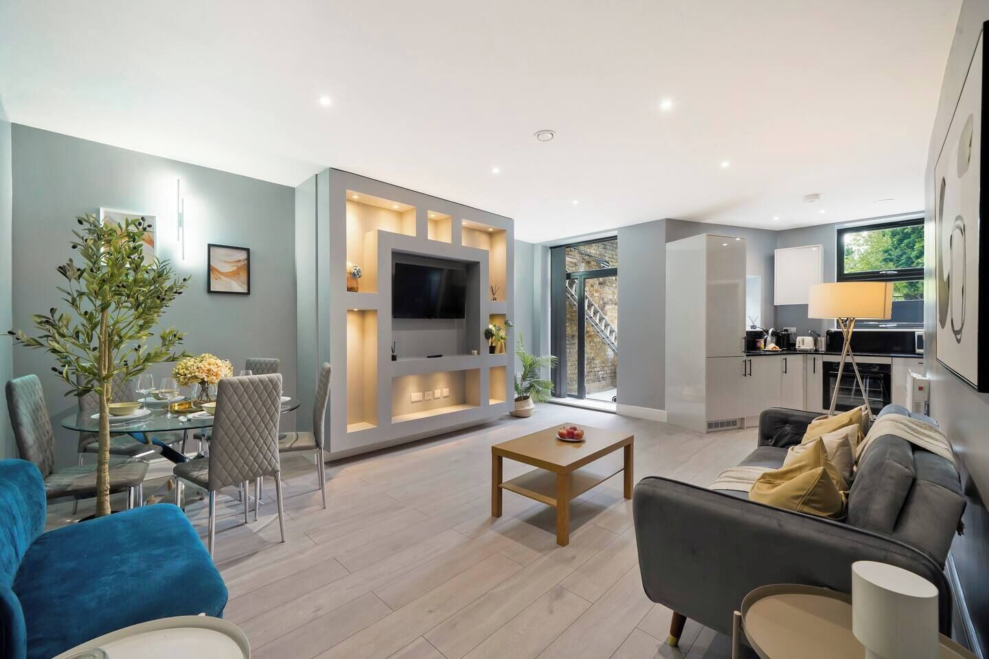 A modern living room with a flat-screen TV, a dining area with a glass table, and a kitchen with a built-in oven.