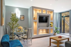 A modern living room with a glass dining table, cushioned chairs, a flat-screen TV, and a staircase leading to an upper level. - 4BR w/ Jacuzzi, Garden & Parking (London)