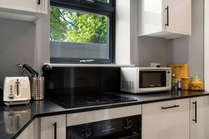 A modern kitchen with a black induction cooktop, a white toaster, a microwave, and a window with a view of greenery. - 4BR w/ Jacuzzi, Garden & Parking (London)