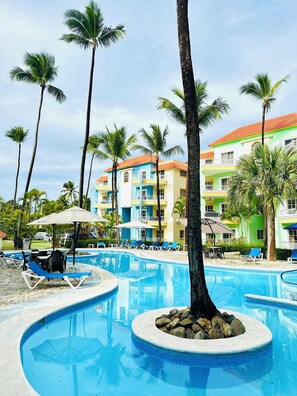 Zona de piscina en un resort con sillas de descanso y sombrillas, rodeada de palmeras y edificios de colores.