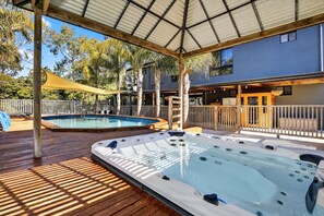 Ein Poolbereich mit zwei Whirlpools, einer Holzterrasse und einer überdachten Sitzgruppe mit Blick auf ein Haus.