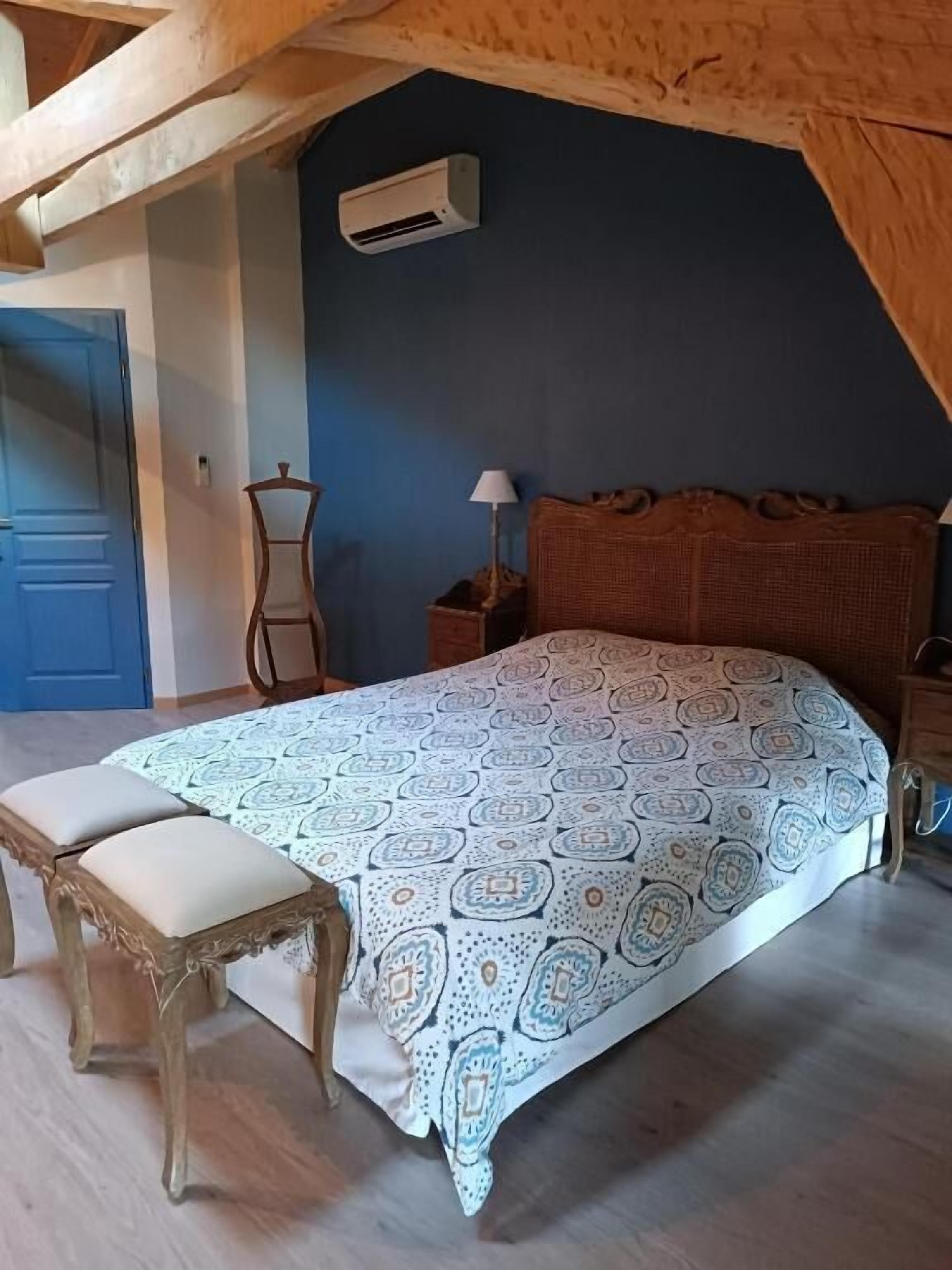 A bedroom with a bed, a bench, a nightstand, a lamp, and an air conditioning unit.