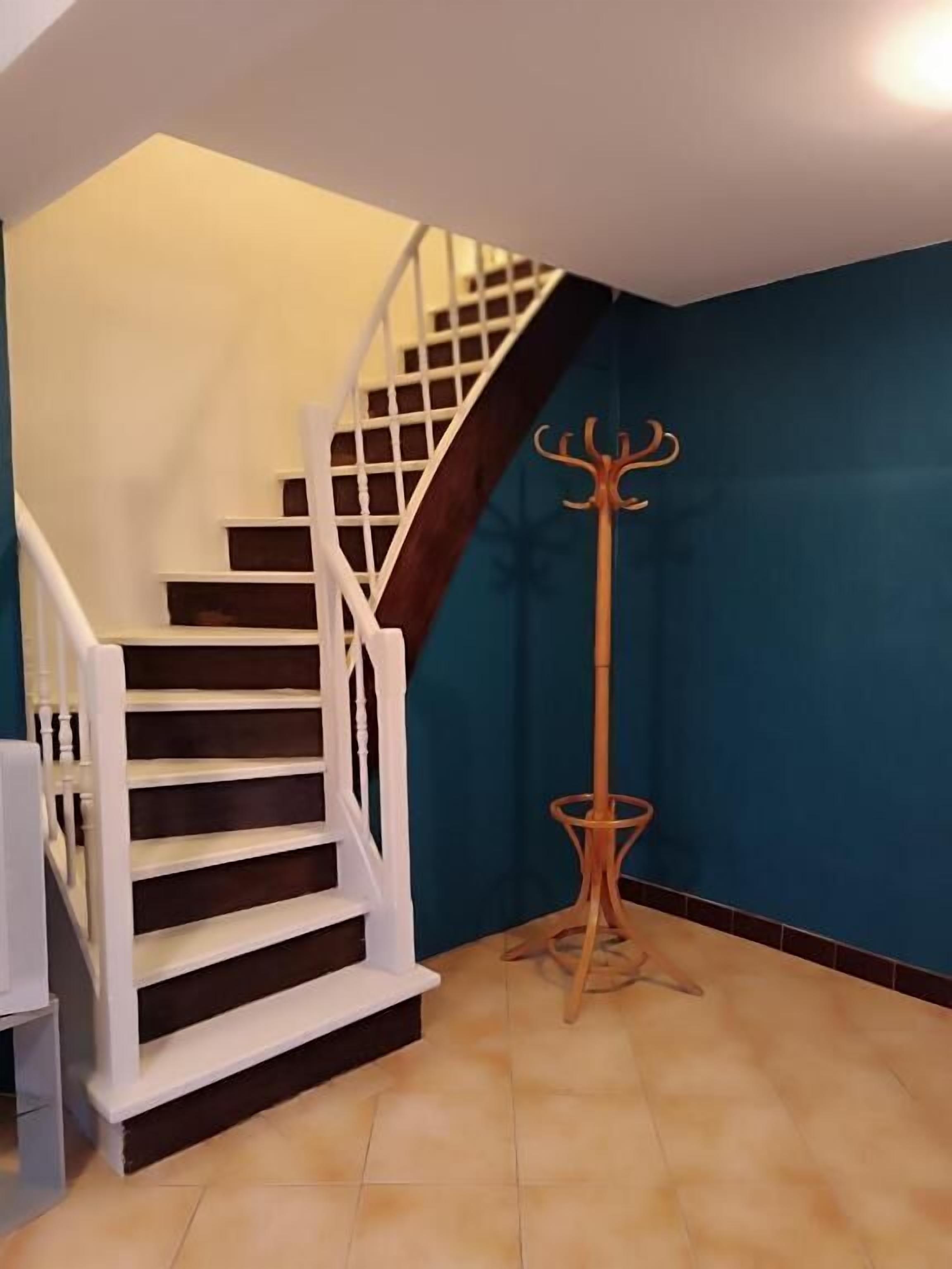 Una escalera con barandillas blancas y peldaños oscuros, que conduce a un rellano con pared azul y un perchero de madera.