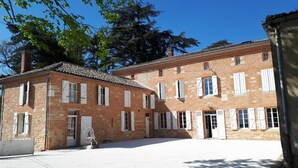 A two-story brick building with a courtyard, featuring multiple windows with shutters, a central door, and a small statue in the foreground. - Le Jardin des Cèdres (Lavaur)