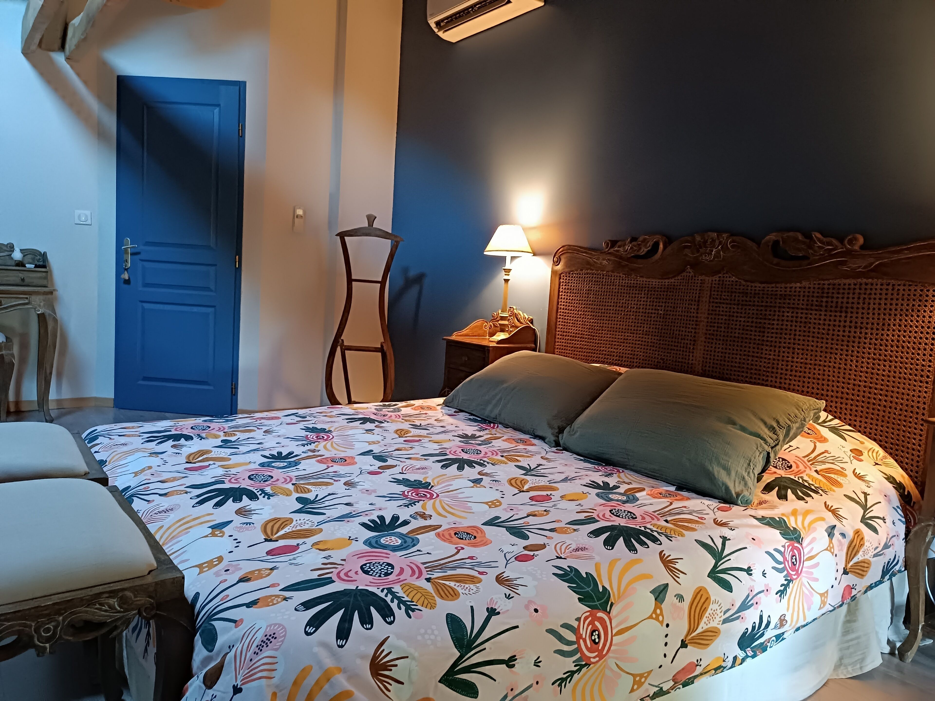 Un dormitorio con colcha estampada de flores, una puerta azul, una mesita de noche con lámpara y un cabecero de ratán.