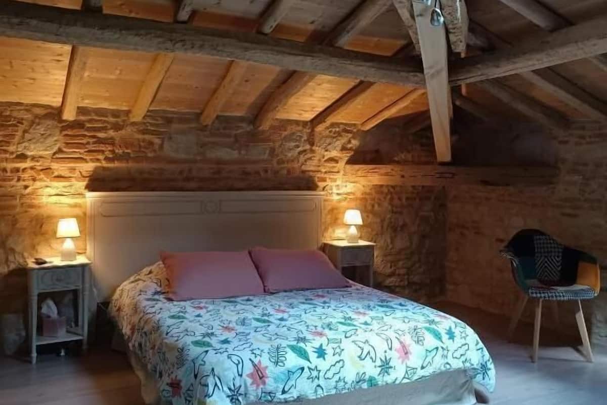 Ein gemütliches Schlafzimmer im Dachgeschoss mit Holzbalkendecke, einem Bett mit floralem Bettzeug, zwei Nachttischlampen und einem Sessel.