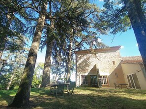 A stone building with a tiled roof, surrounded by trees and a grassy area with outdoor furniture. - Le Jardin des Cèdres (Lavaur)