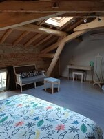 Un dormitorio tipo loft con techo inclinado, cama con colcha floral de colores, un banco, una mesita y una silla.