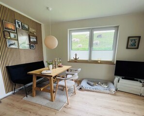 Una habitación acogedora con una mesa de comedor de madera, un sofá negro, un televisor y una ventana que da a ovejas pastando.