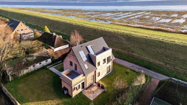 A modern two-story house with a large patio area, situated in a rural setting with a view of a water body and wind turbines in the distance. - Deichkoje12 - Deck 1 vacation apartment directly on the dike with dog | North Sea near (Tönning)
