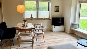 A modern living room with a wooden dining table, four chairs, a television, and a window with blinds. - Deichkoje12 - Deck 1 vacation apartment directly on the dike with dog | North Sea near (Tönning)