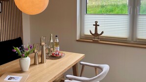 A dining area with a wooden table, white chairs, a hanging light fixture, and a window with a view of sheep grazing. - Deichkoje12 - Deck 1 vacation apartment directly on the dike with dog | North Sea near (Tönning)
