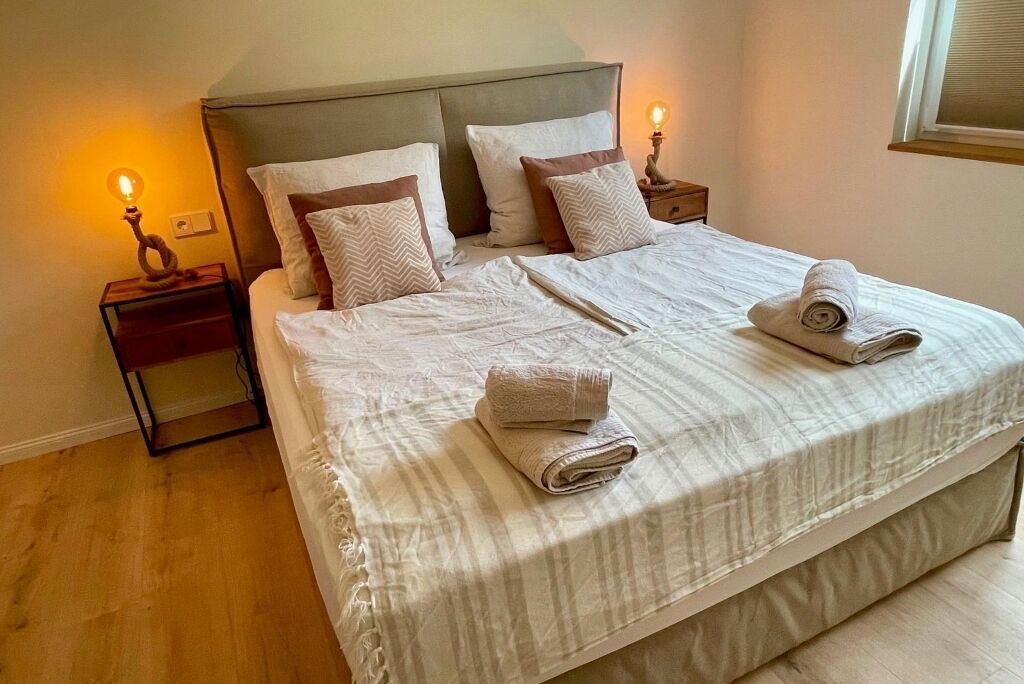 A neatly made bed with a striped bedspread, two pillows, and two rolled towels on the bed. There are two bedside tables with lamps, one on each side of the bed.