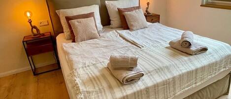 A neatly made bed with a striped bedspread, two pillows, and two rolled towels on the bed. There are two bedside tables with lamps, one on each side of the bed.