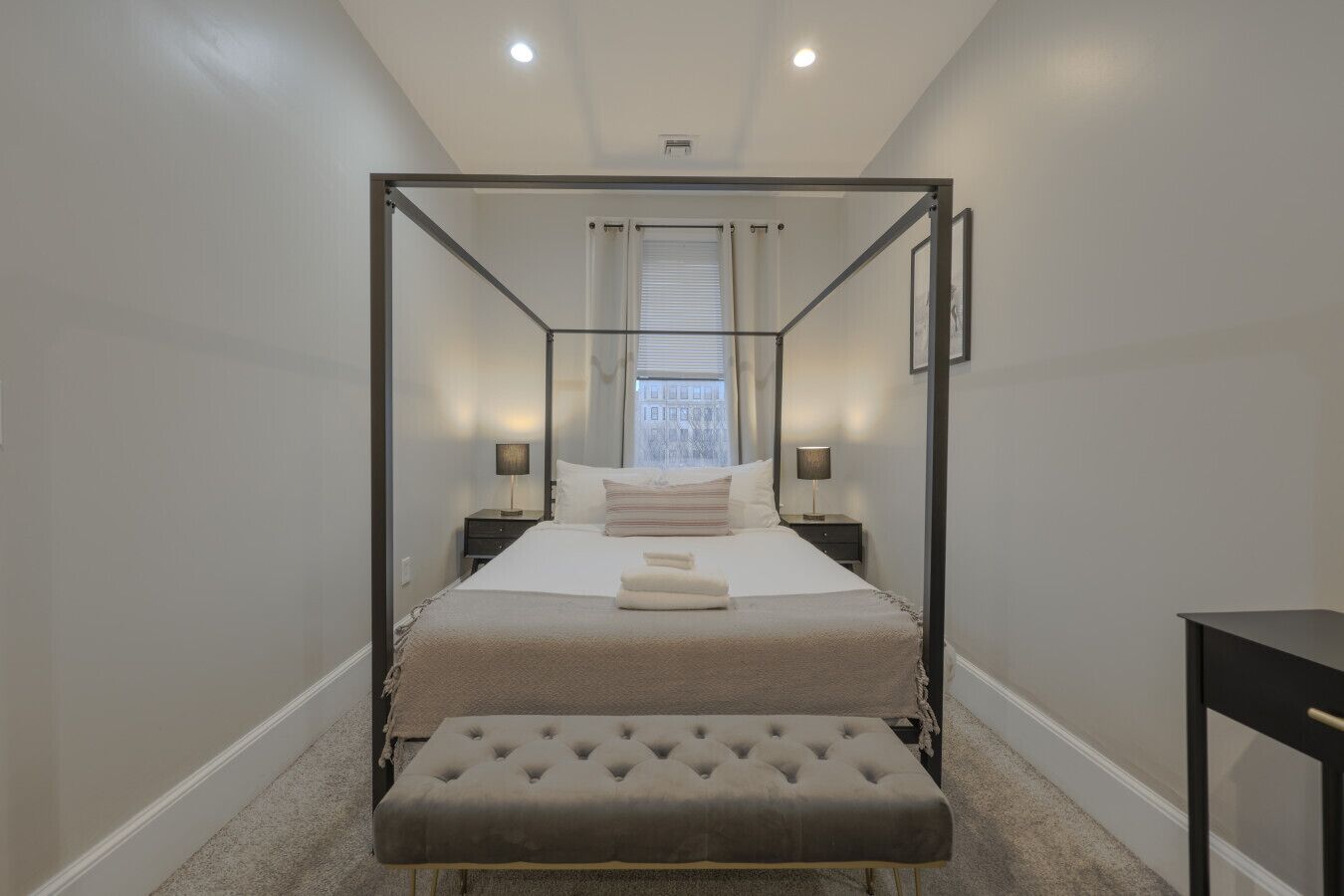 A four-poster bed with a tufted ottoman, bedside tables with lamps, and a view of the outside through a window with blinds.