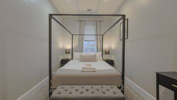 A four-poster bed with a tufted ottoman, bedside tables with lamps, and a view of the outside through a window with blinds.