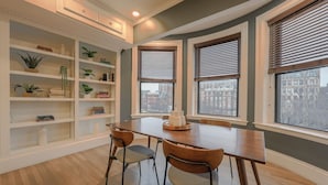 A dining area with a wooden table and chairs, a white built-in bookshelf, and three windows with blinds. - Luxury Home located in Back Bay 8min walk to Fewnway Park, close to Boston University, Berklee & MIT (Boston)