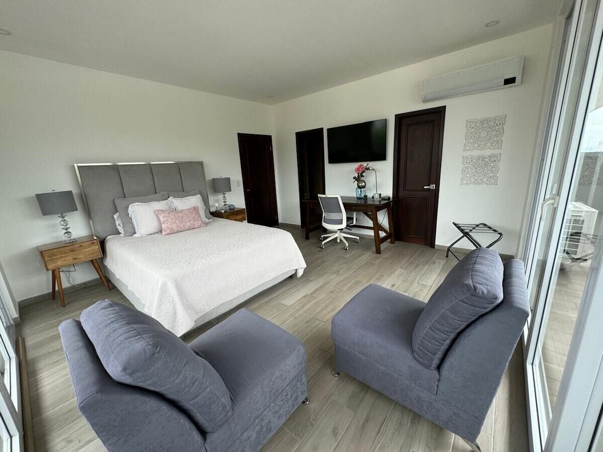 Ett modernt hotellrum med en stor säng, två grå fåtöljer, ett skrivbord med en stol, en TV och ett fönster med utsikt.