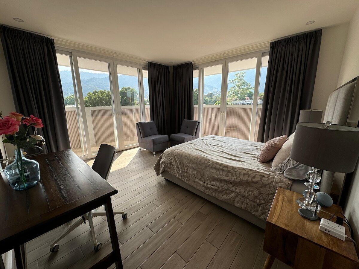 Ein Hotelzimmer mit einem großen Bett, einem Schreibtisch mit Blumenvase, zwei Stühlen, einer Lampe und Blick ins Freie.
