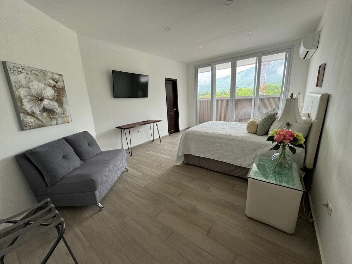 A bedroom with a bed, a grey sofa, a small table with a vase, a TV on the wall, and a window with a view of greenery.