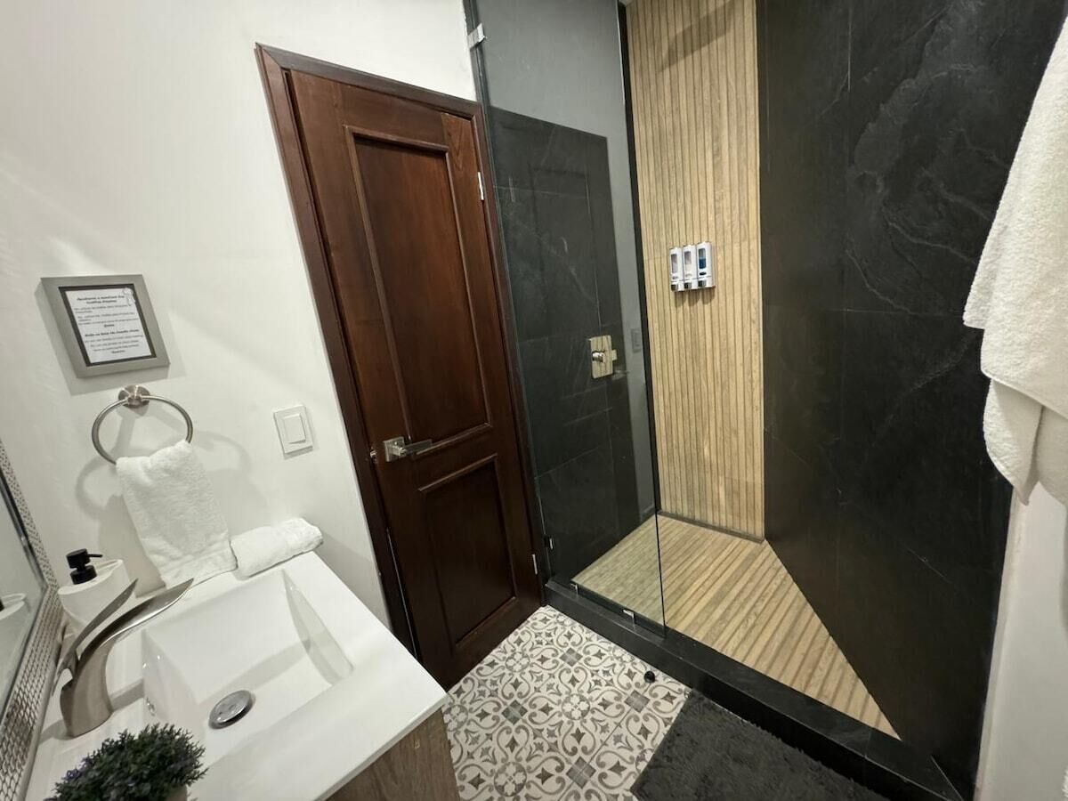 Een badkamer met een houten deur, een glazen douchecabine, een wit wastafelmeubel en een vloerkleed met dessin.