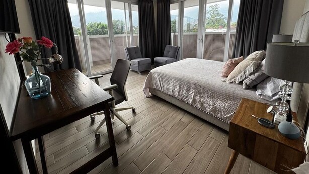 A hotel room with a bed, a desk, a chair, a small table with a lamp, and a vase with flowers. - Luxury 3BR Penthouse City Mall SPS (San Pedro Sula)