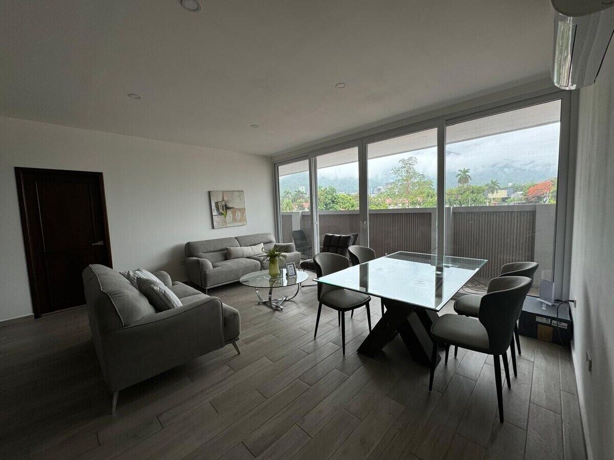 A modern living room with a glass dining table, grey sofas, and a large window offering a view of greenery and mountains.