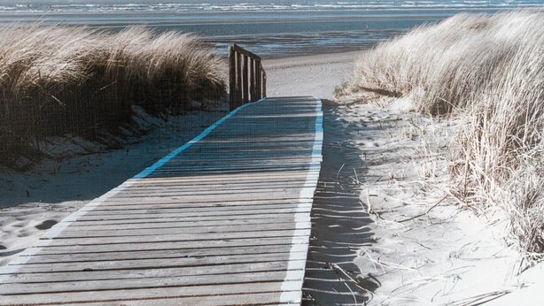 Ein hölzerner Steg führt zum Strand, begleitet von Dünen auf beiden Seiten.