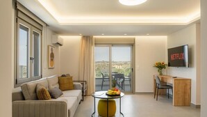 Een moderne woonkamer met een bank, salontafel en een bureau met stoel. Een televisie is aan de muur bevestigd, en er is een balkon zichtbaar door de glazen deuren.