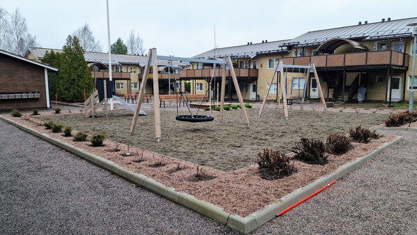 A playground with swings and a tire swing, surrounded by a gravel area with small shrubs.