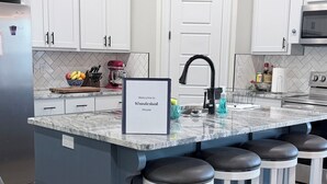 Una cocina moderna con armarios blancos, encimera de mármol, una isla integrada con taburetes de bar y un refrigerador de acero inoxidable.