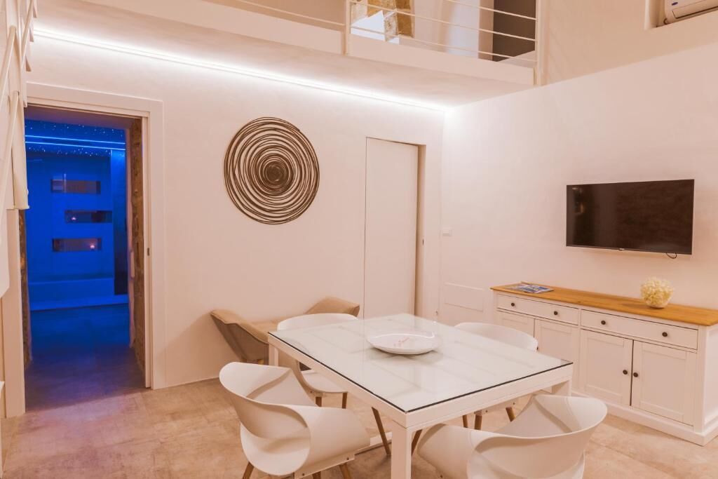 A modern dining area with a glass table, white chairs, a decorative wall piece, a TV, and a wooden sideboard.