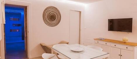 Un moderno comedor con una mesa de vidrio, sillas blancas, un adorno en la pared, un televisor y un aparador de madera.