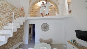 Un interior moderno con pared de piedra, una escalera blanca, una zona de comedor y un televisor sobre un mueble de madera.