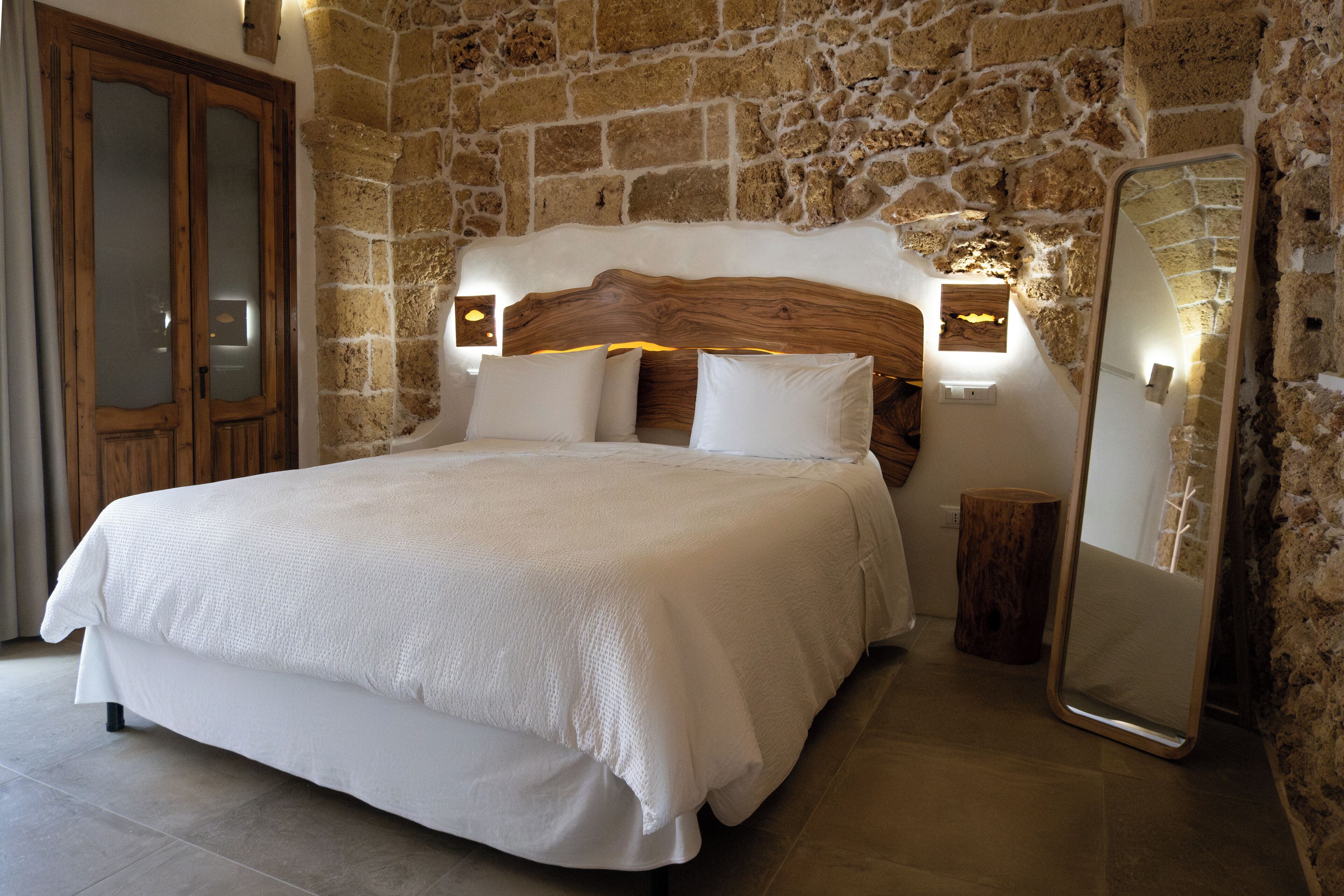 A bedroom with a stone wall, a wooden headboard, a bed with white bedding, a wooden nightstand, and a mirror.
