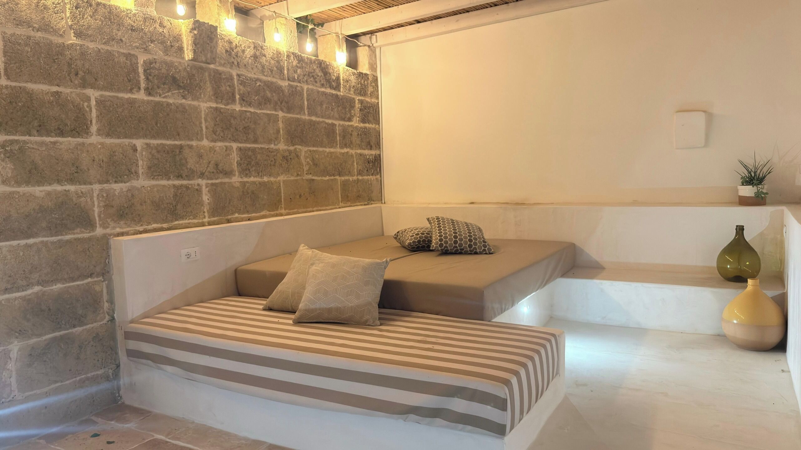 A cozy room with a stone wall, a bed with striped bedding, and a small seating area with cushions.