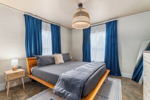 Ein Schlafzimmer mit einem Bett, einem Nachttisch mit Lampe und einem großen Fenster mit Vorhängen.