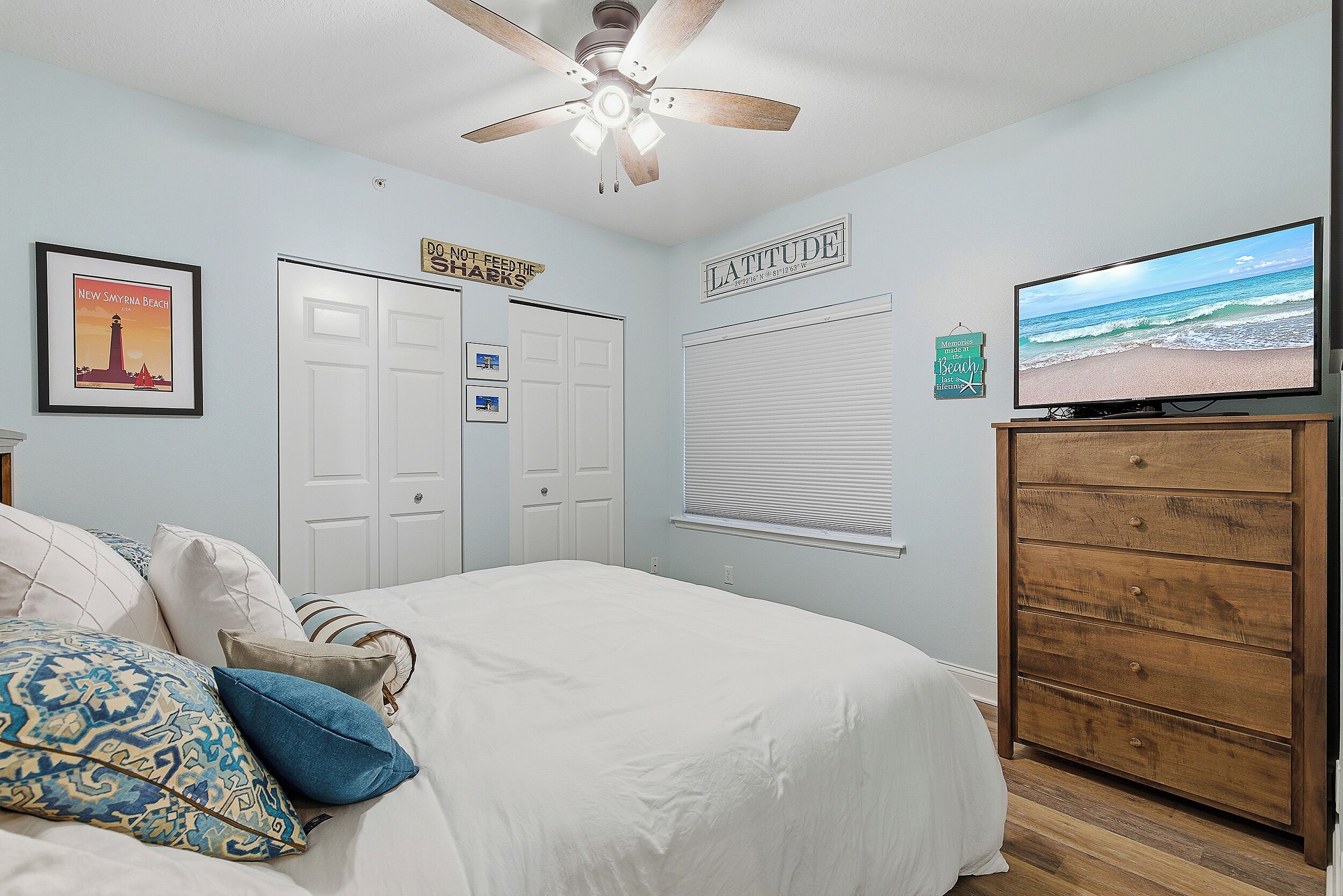 Quarto com cama, ventilador de teto, televisão, cômoda e parede com pôster de praia e um letreiro.