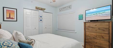 Quarto com cama, ventilador de teto, televisão, cômoda e parede com pôster de praia e um letreiro.