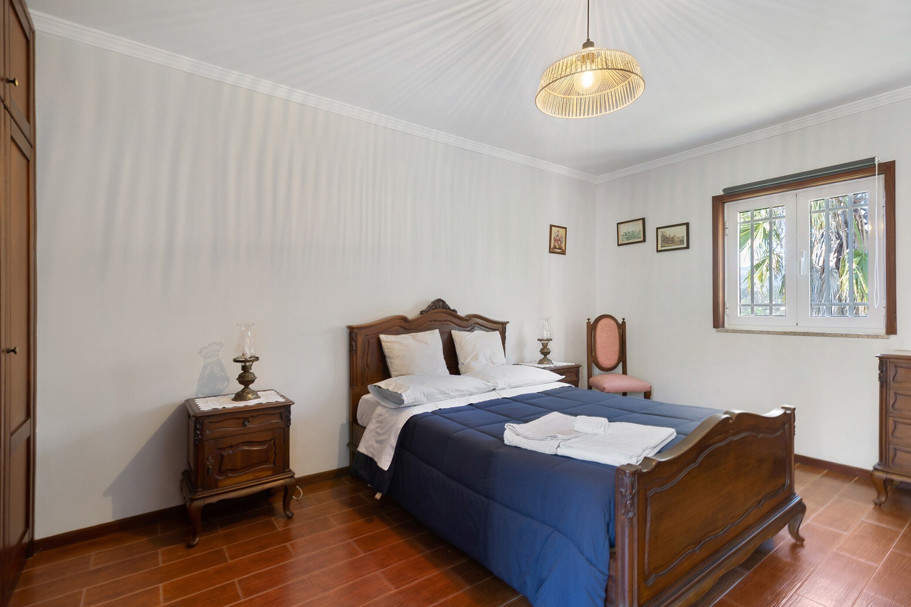 A bedroom with a wooden bed, a nightstand, a dresser, and a window with a view of palm trees.
