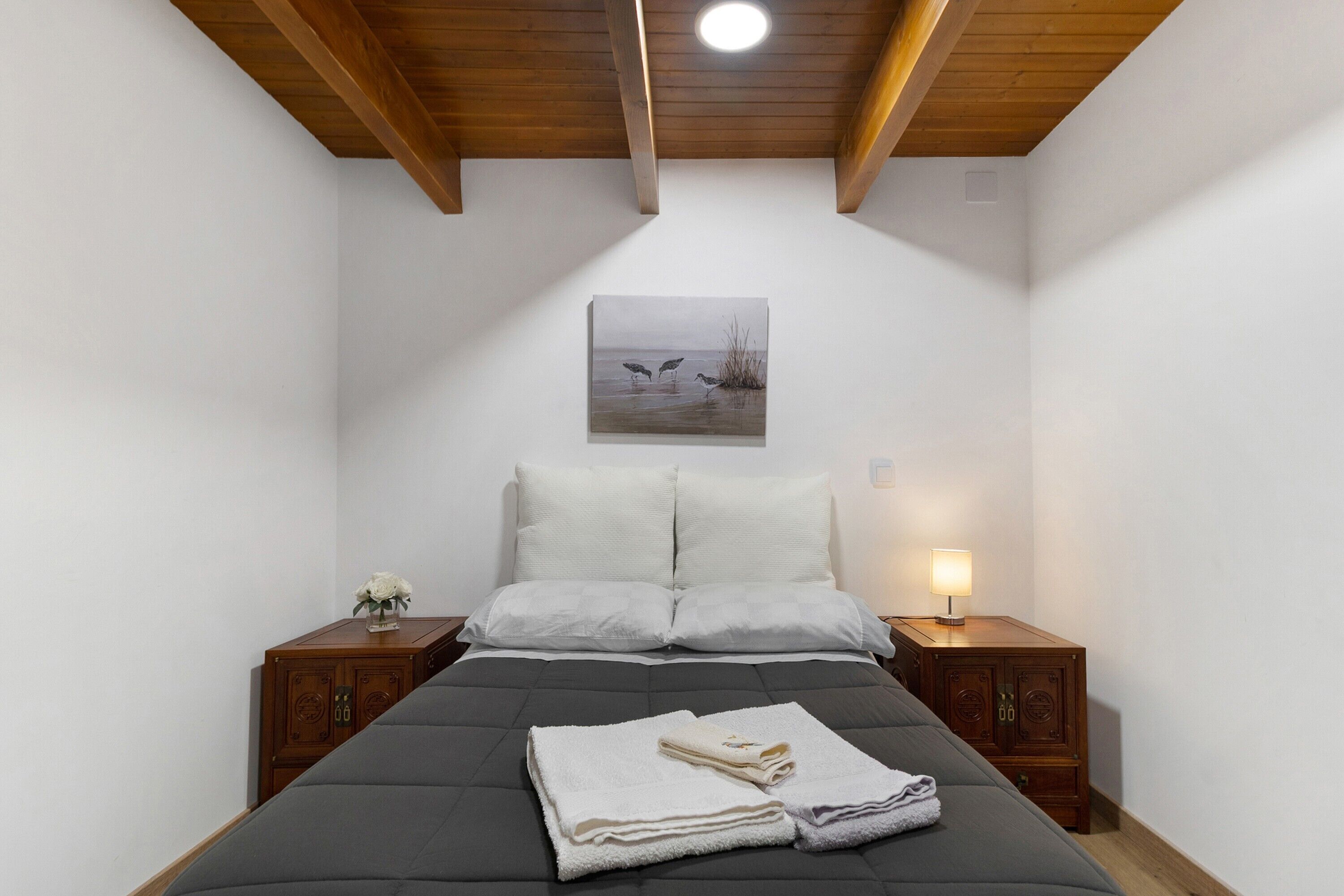 A bedroom with a bed, two nightstands, a night lamp, a painting, and a wooden ceiling.