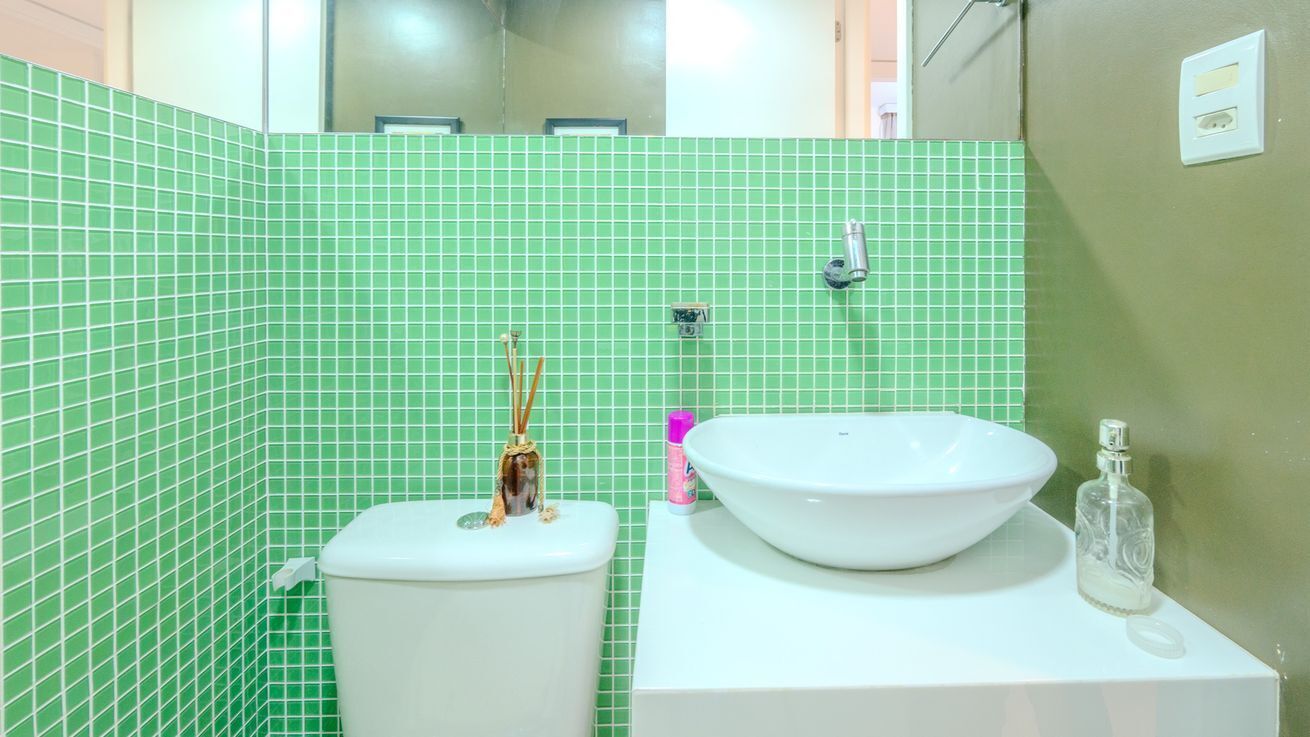 Un baño con paredes de azulejos verdes, un inodoro blanco y un lavabo blanco con un recipiente grande, además de una pequeña botella de jabón líquido.