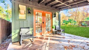 A covered patio with a wooden deck, two wicker chairs, a red door, and a view of a grassy yard. - Loft Jason | Relaxing Lake Conroe Getaway (Montgomery)