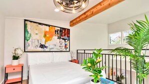 A bedroom with a large bed, a painting, a nightstand, a potted plant, and a view of the outdoors. - Loft Jason | Relaxing Lake Conroe Getaway (Montgomery)