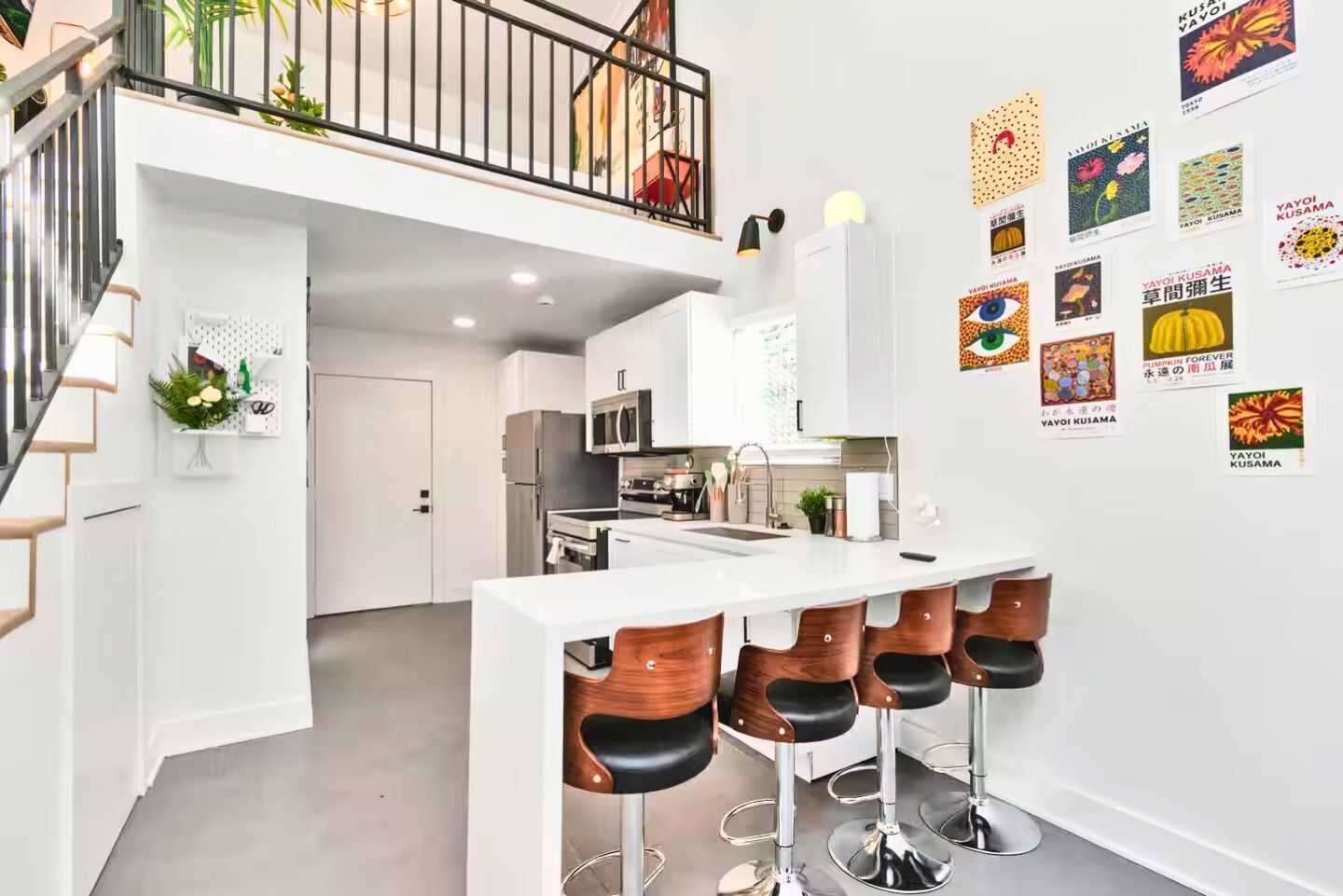 A modern kitchen with a central island and bar stools, a staircase, and a wall decorated with posters.