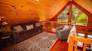 A cozy wooden cabin interior with a loft bedroom, a living room with a sofa and a chair, and a large window offering a view of the outdoors. - Anisidi | New River Waterfront • Pet Friendly (Pulaski County)