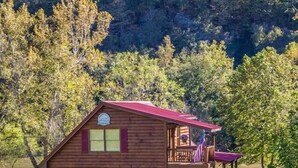 A two-story log cabin with a balcony, surrounded by a grassy area and trees. - Anisidi | New River Waterfront • Pet Friendly (Pulaski County)