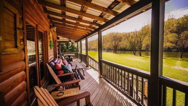 A wooden cabin with a porch featuring a sofa and chairs, a table, and a view of a grassy area with trees. - Anisidi | New River Waterfront • Pet Friendly (Pulaski County)