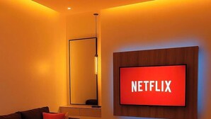 Ein modernes Wohnzimmer mit einer Couch, einem Couchtisch mit Früchten und Gläsern sowie einem an der Wand befestigten Fernseher, der "NETFLIX" anzeigt.